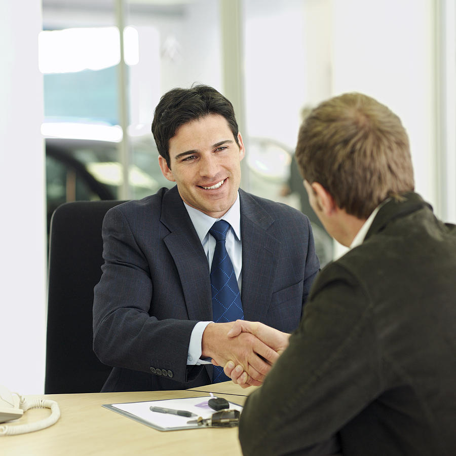 Car salesman shaking hands over desk with customer in car showroom Photograph by Stockbyte