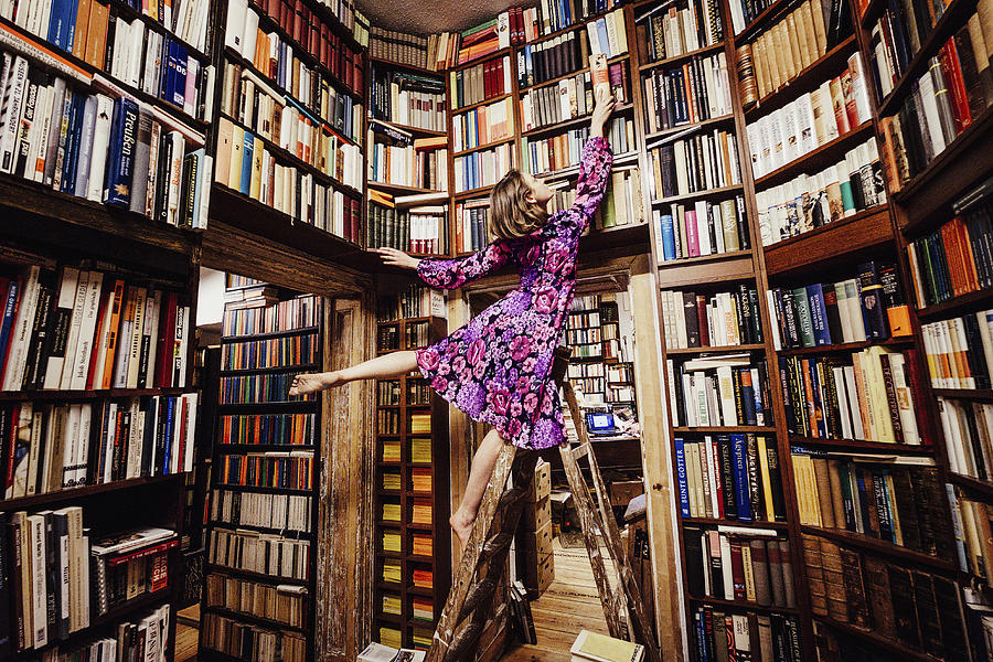 Carefree woman on ladder reaching for book in library Photograph by Sven Hagolani