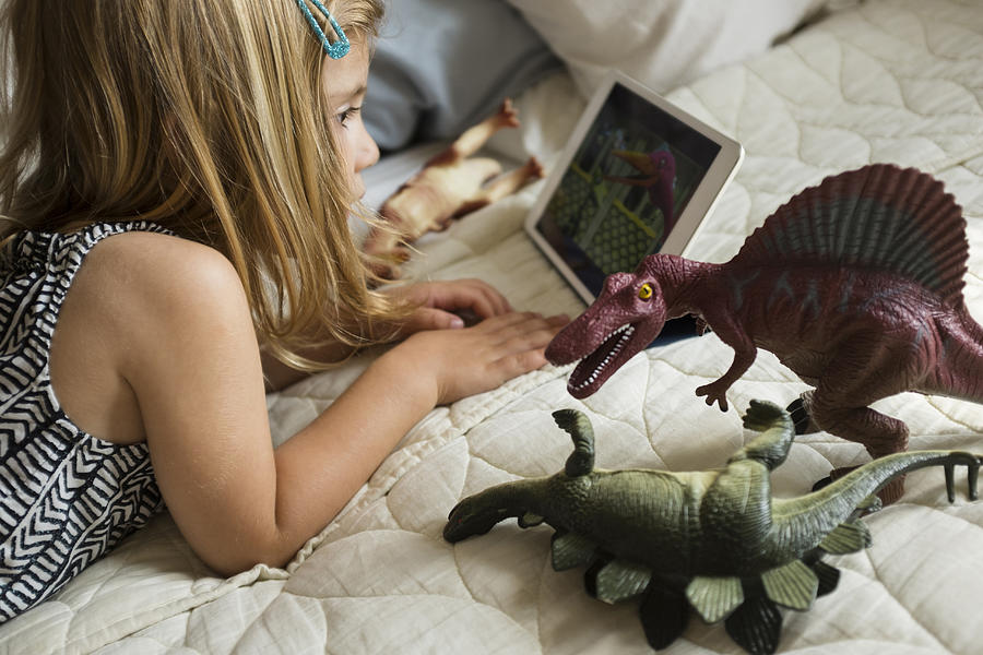 Caucasian girl laying on bed with toy dinosaurs using digital tablet Photograph by JGI/Jamie Grill