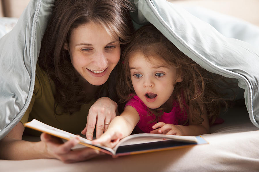 Caucasian mother and daughter reading book under covers Photograph by Mike Kemp