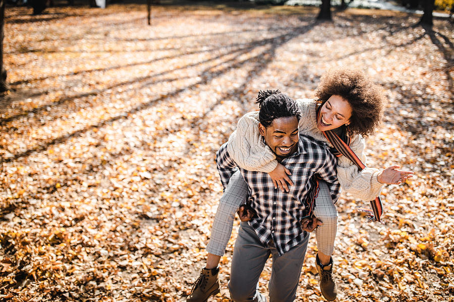 Cheerful African American couple piggybacking in autumn at the park. Photograph by Skynesher