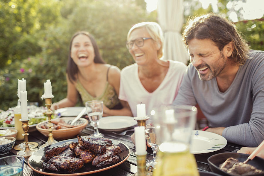 Cheerful couple and female friend laughing on dining table during garden party in back yard Photograph by Maskot