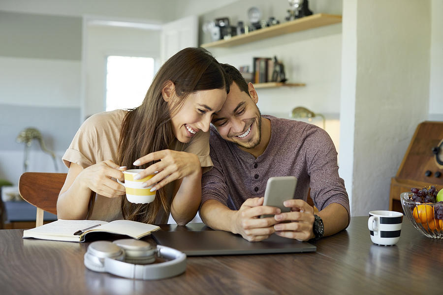 Cheerful couple using mobile phone in dining room Photograph by Morsa Images