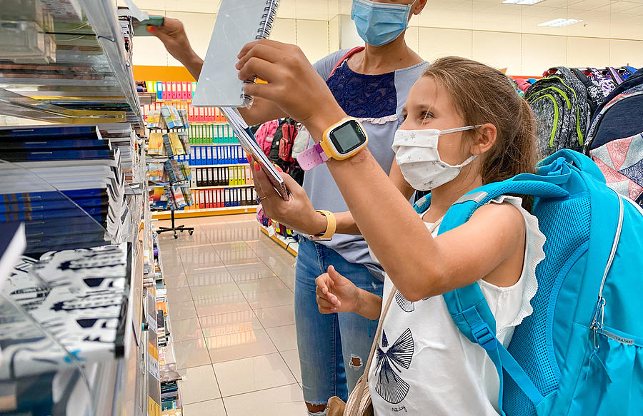 Cheerful Girl Buying Workbooks on Back To School Shopping with her Mother Wearing Protective Face Mask - stock photo Photograph by CasarsaGuru