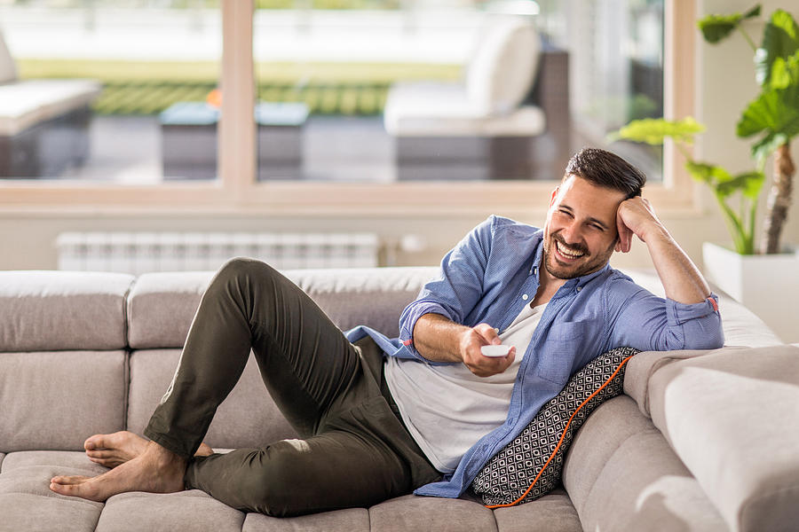 Cheerful man changing channels while relaxing on sofa at home. Photograph by Skynesher