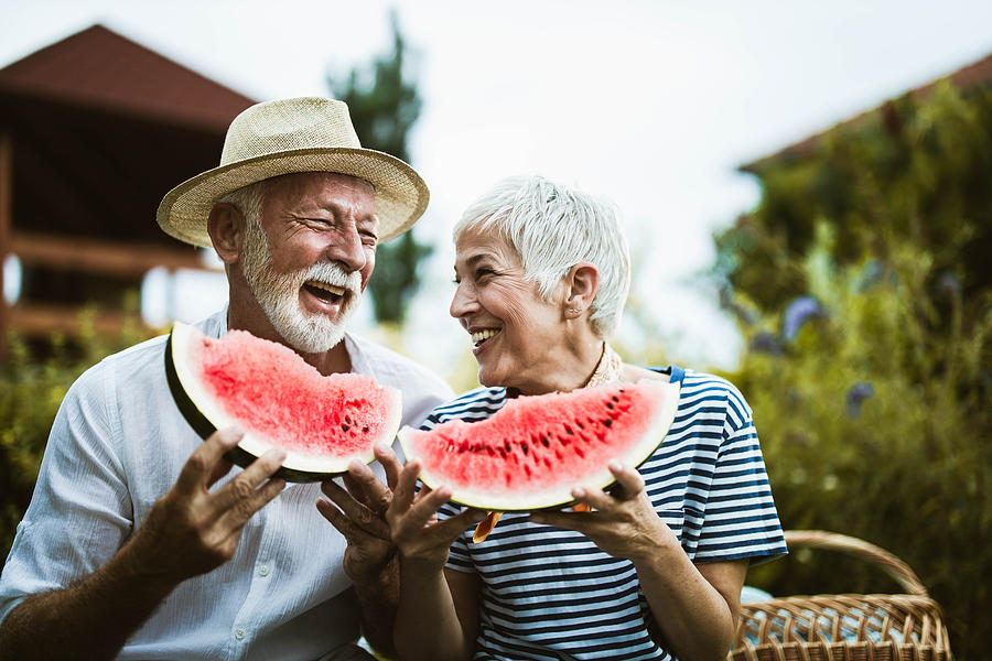 Cheerful mature couple having fun while eating watermelon during picnic day in nature. Photograph by Skynesher