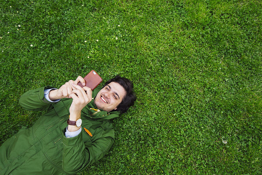 Cheerful smiling young man lying on grass with smartphone Photograph by Alexander Spatari