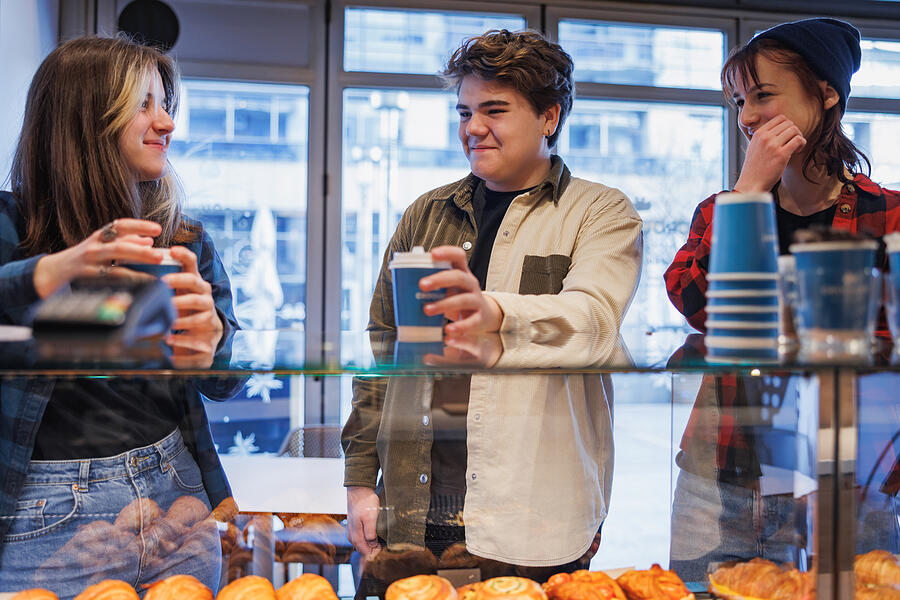 Cheerful teenagers standing by the baked pastry showcase counter, having coffee in paper cups Photograph by Zoranm