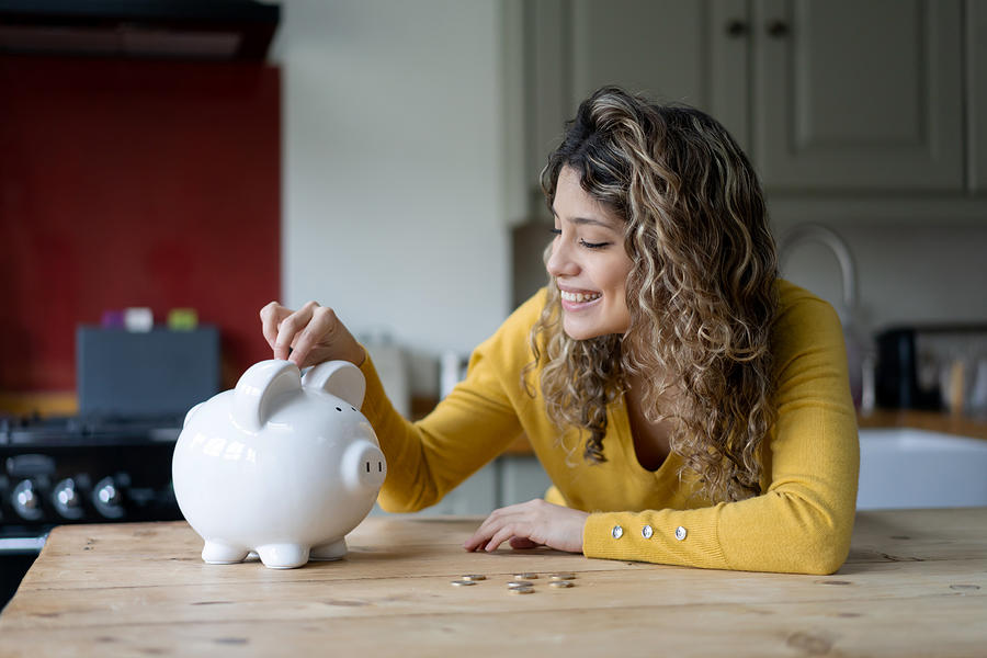 Cheerful young woman with curly hair at home saving coins into her piggybank Photograph by Hispanolistic