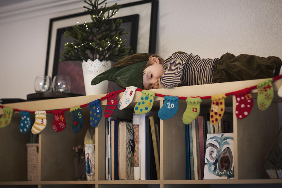 Child (6-7) dressed as an elf, sleeping on top of a bookshelf Photograph by Elva Etienne
