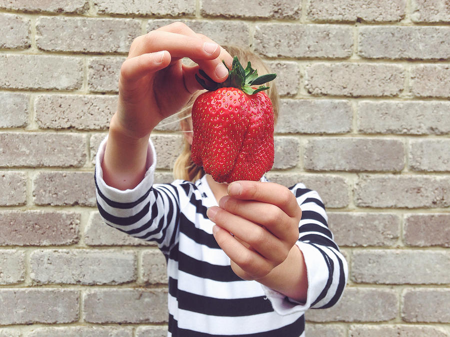 Child holding large strawberry in front of face Photograph by Jodie Griggs