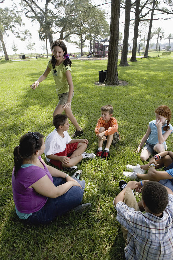 Children sitting in a circle on the grass with their teacher while a girl walks around them Photograph by Purestock