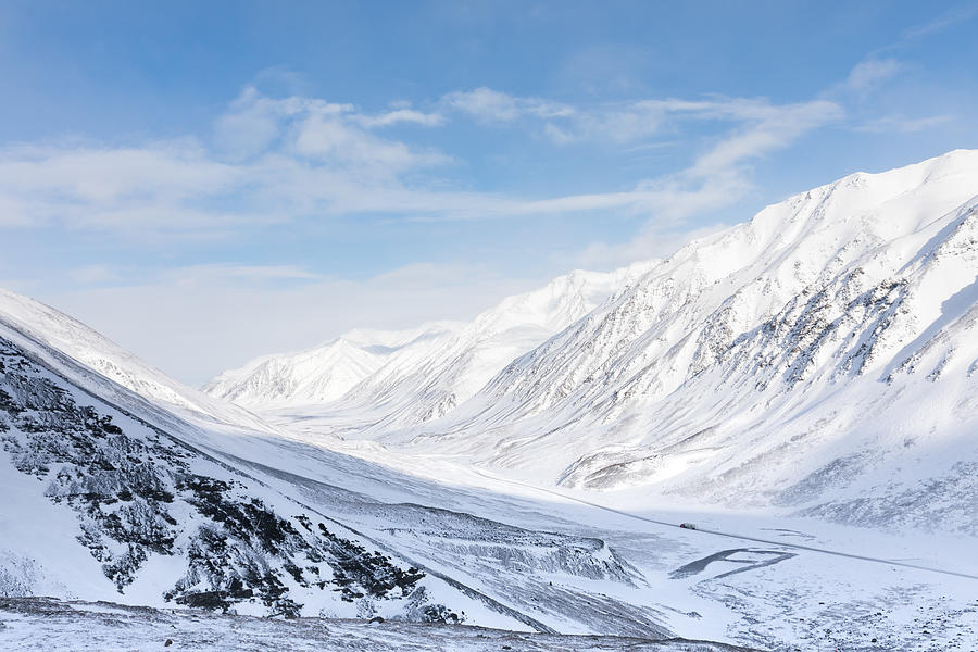 Clear winters day over the scenic snowy alaskan mountain range with the sun highlighting the peaks. A transport truck winds it way down through the valley on the dalton highway. Photograph by Diane Keough