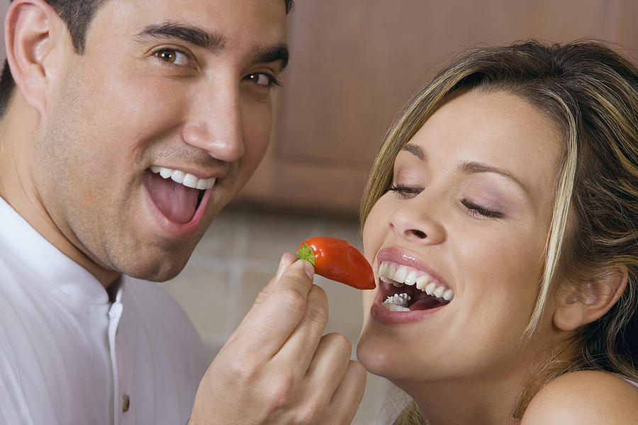 Close-up of a mid adult man feeding a red chili pepper to a young woman Photograph by Glowimages