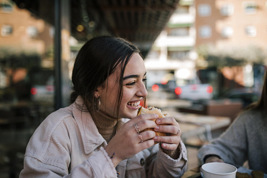 Close-up of cheerful teenage girl eating burger with friend at sidewalk cafe Photograph by Westend61