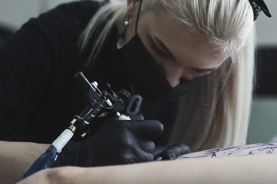 Close-up of female artist wearing mask tattooing on womans thigh with tattoo machine Photograph by Alexandr Sherstobitov