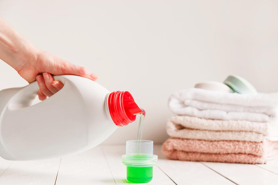 Close up of female hands pouring liquid laundry detergent into cap on white rustic table with towels on background in bathroom. Photograph by Vladdeep