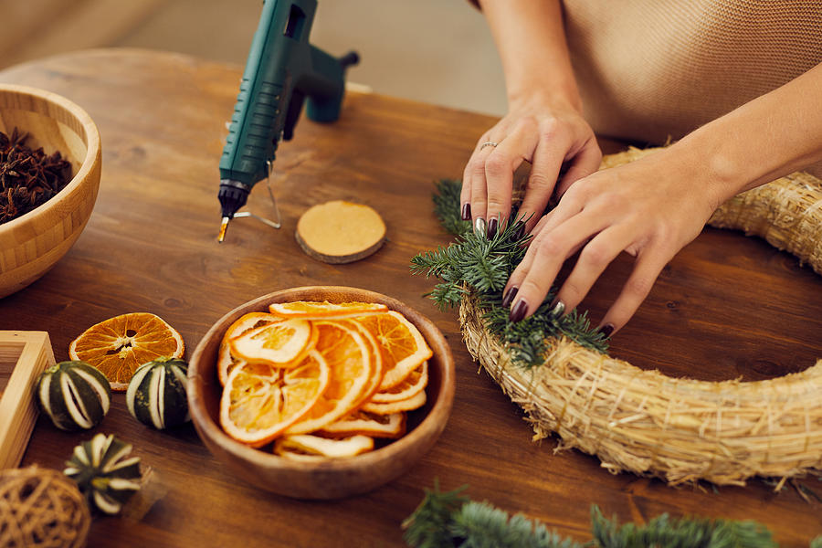 Close-up of unrecognizable woman with manicure standing at wooden table in workshop and using hot glue while attaching fir tree twigs to wreath base Photograph by Mediaphotos