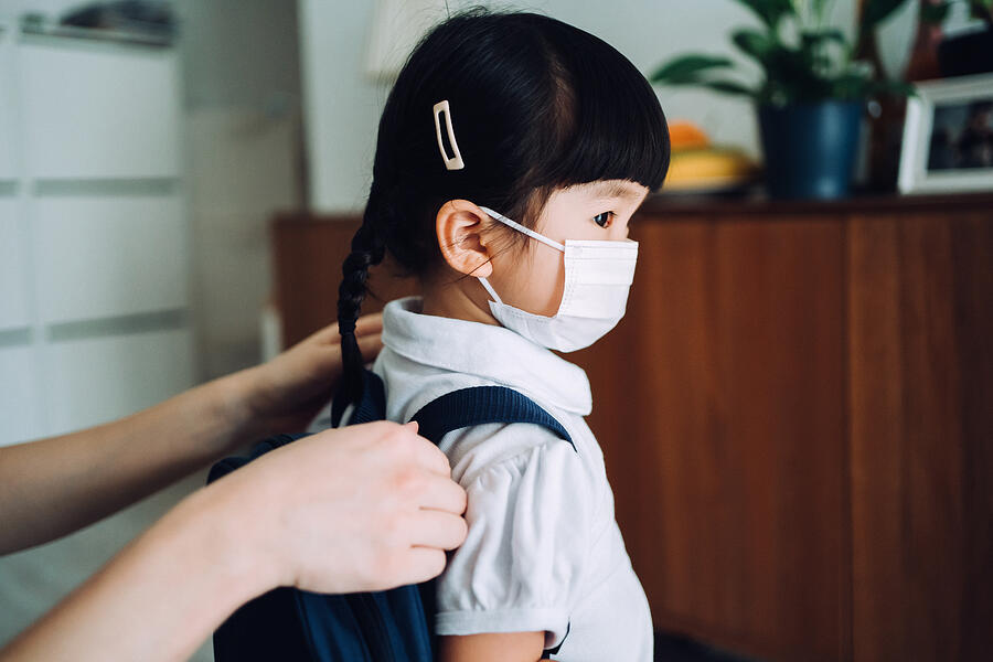 Close up of young Asian mother putting on school satchel for her little daughter in the living room and preparing for her first day at school during the Covid-19 pandemic Photograph by D3sign