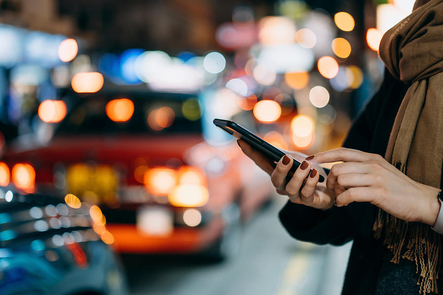 Close up of young woman using mobile app device on smartphone to arrange taxi ride in downtown city street, with illuminated busy city traffic scene during rush hour with traffic congestion in the evening Photograph by D3sign