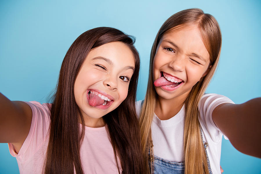 Close up photo two pretty little age girls holiday having fun funky tongue out of mouth make take selfies long pretty hair wearing casual jeans denim t-shirts isolated on blue bright background Photograph by Deagreez
