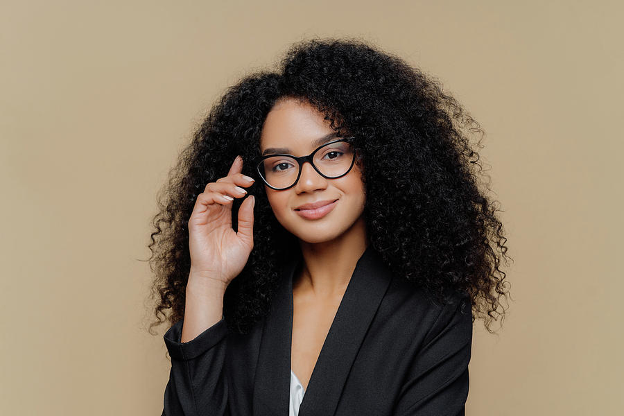 Close up portrait of calm satisfied Afro American woman with bushy curly  hairstyle, keeps hand raised, has healthy skin, wears spectacles, black  elegant outfit isolated on brown wall. Face expressions Photograph by