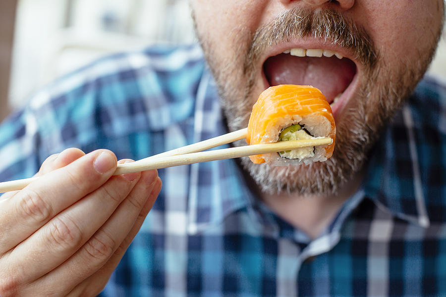 Close up view of man with beard eating sushi roll with salmon using chopsticks Photograph by Alexander Spatari