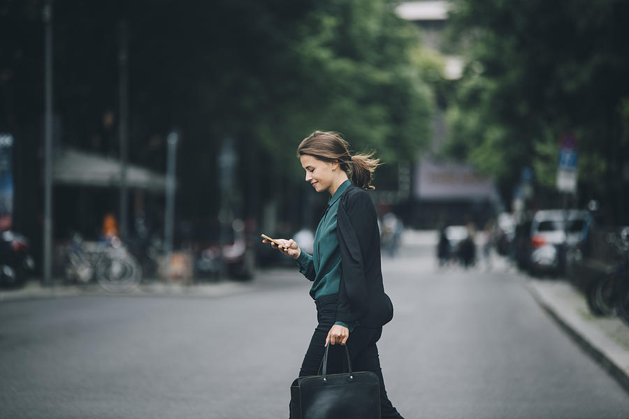 Confident businesswoman using smart phone while crossing street in city Photograph by Maskot