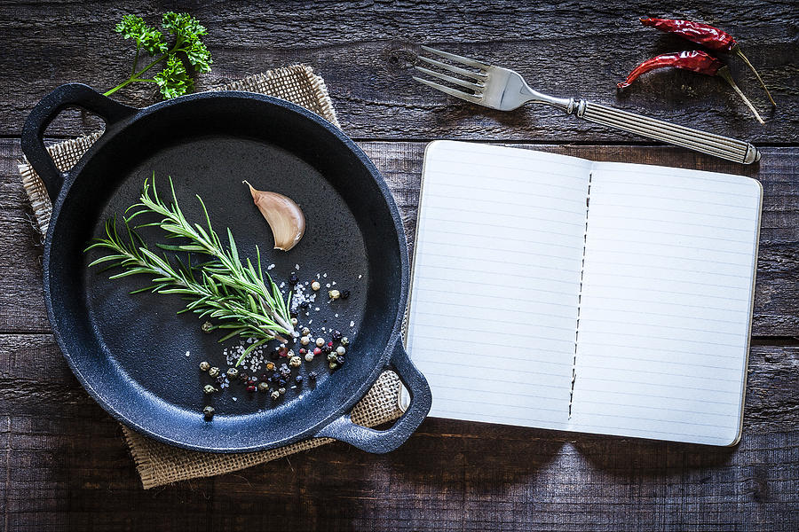 Cookbook and cast iron pan with some herbs on wooden table Photograph by Fcafotodigital