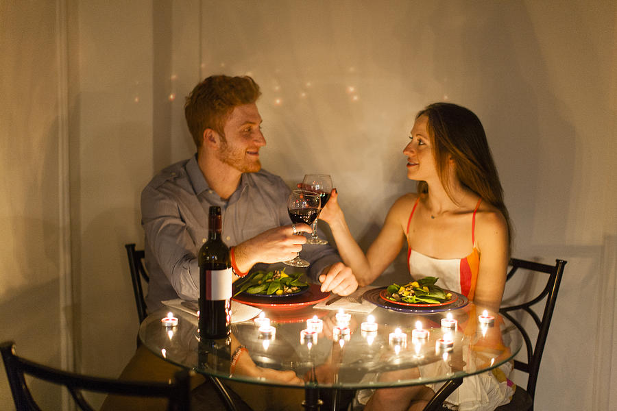 Couple at table face to face enjoying candlelit meal, making a toast, smiling Photograph by Zave Smith