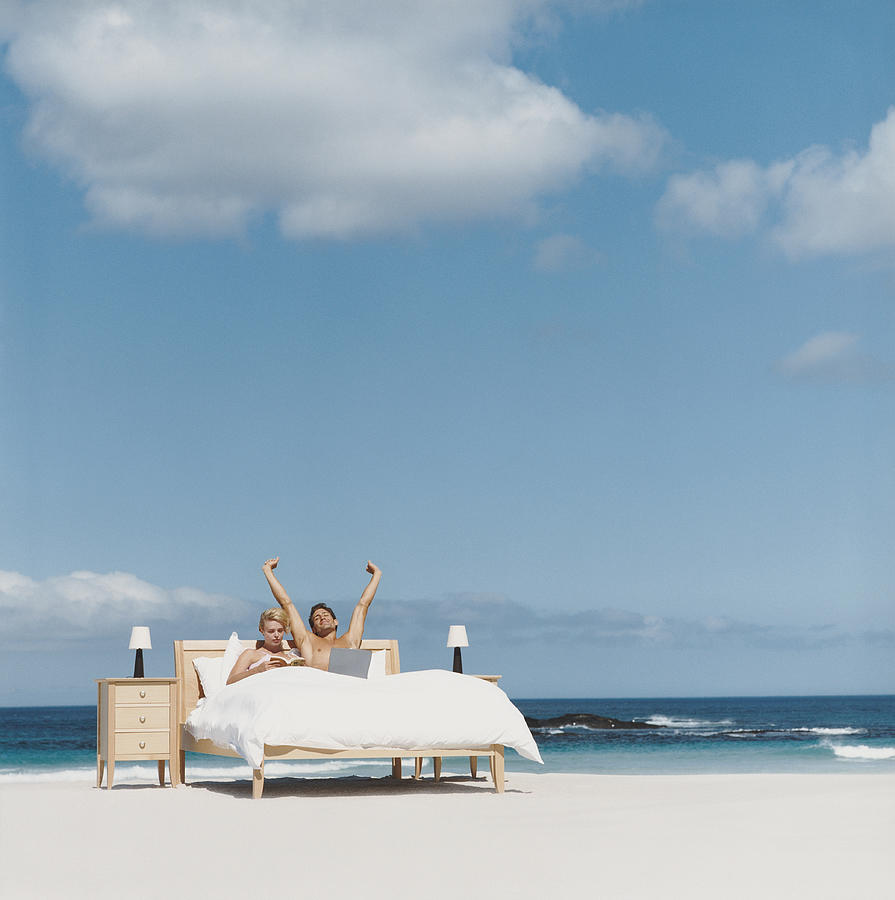 Couple in Bed on a Beach, Man Stretching His Arms Photograph by Digital Vision.