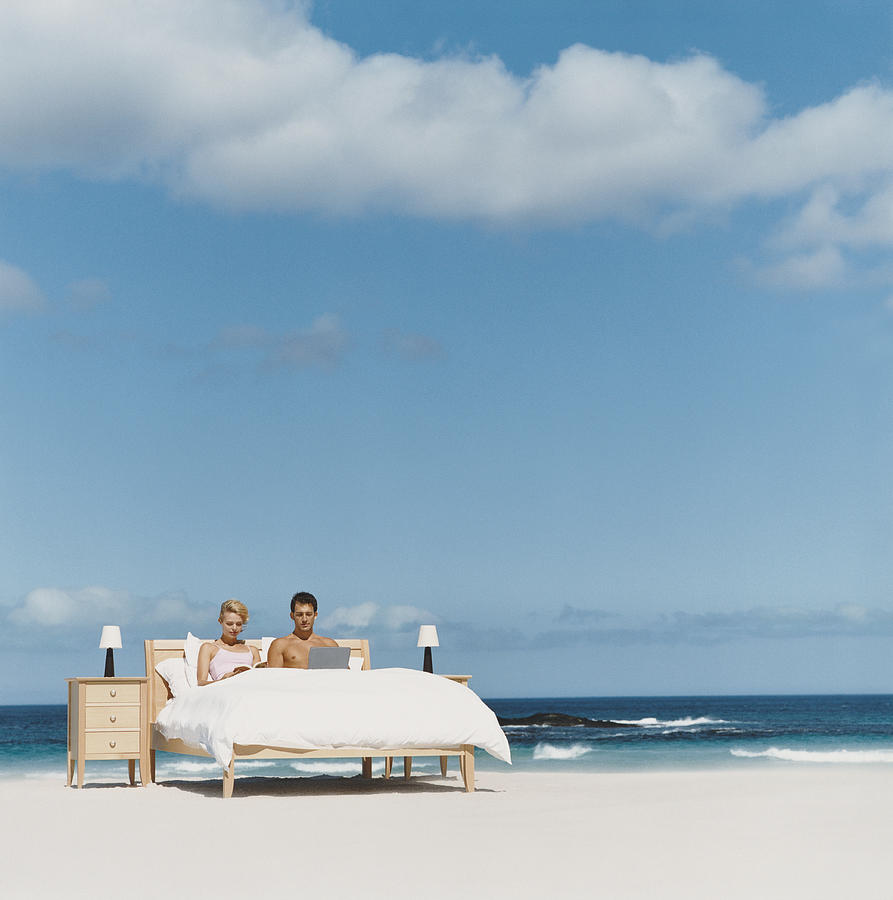 Couple Lying in Bed on a Beach, Man Using His Laptop and Woman Reading a Book Photograph by Digital Vision.