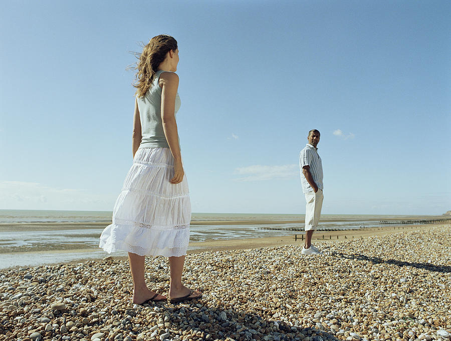 Couple on pebble beach, woman looking at man with hands in pockets Photograph by Nick White