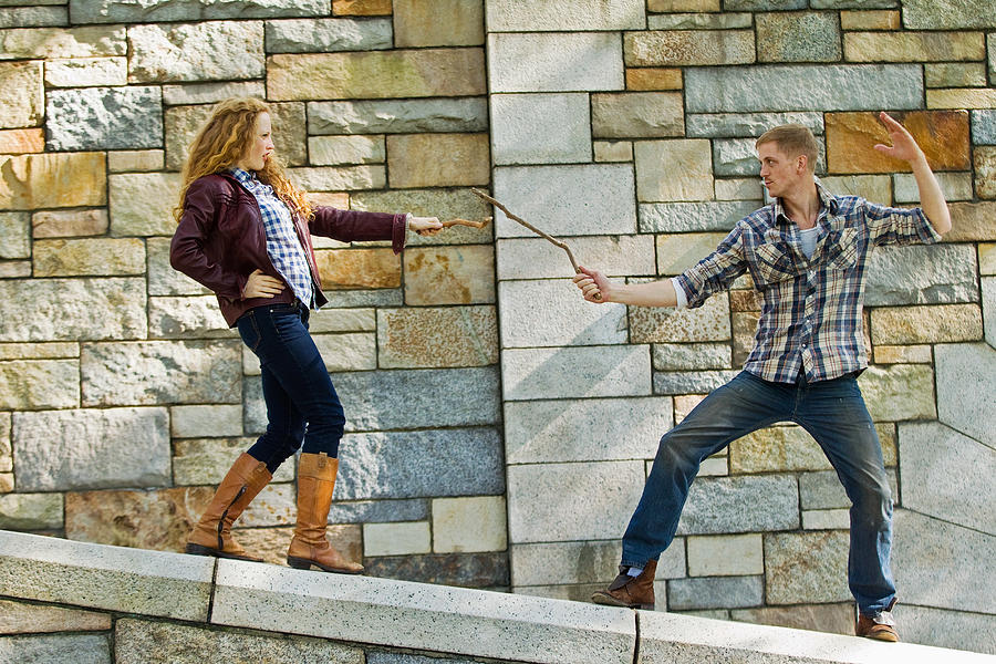 Couple play fighting with sticks against stone wall Photograph by Image Source