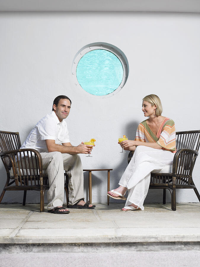 Couple Sit Holding Cocktails on a Pavement in Front of a Wall With a Port-Hole Photograph by Digital Vision.