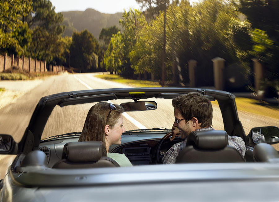 Couple Sit in the Front Seat of a Convertible, Driving on the Road and Smiling at Each Other Photograph by Digital Vision.