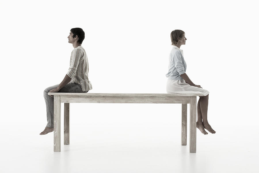 Couple sitting back to back on wooden table against white background, side view Photograph by Knowlesie