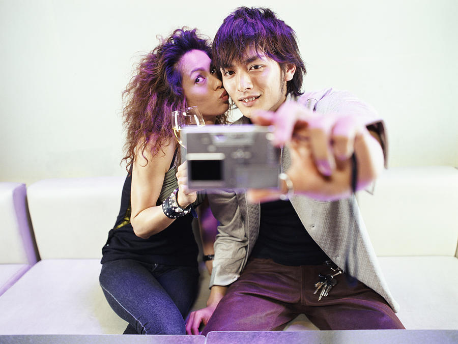 Couple Sitting on a Sofa, Man Taking a Photograph of Themselves With a Digital camera Whilst The Woman is Kissing His Cheek Photograph by Digital Vision.