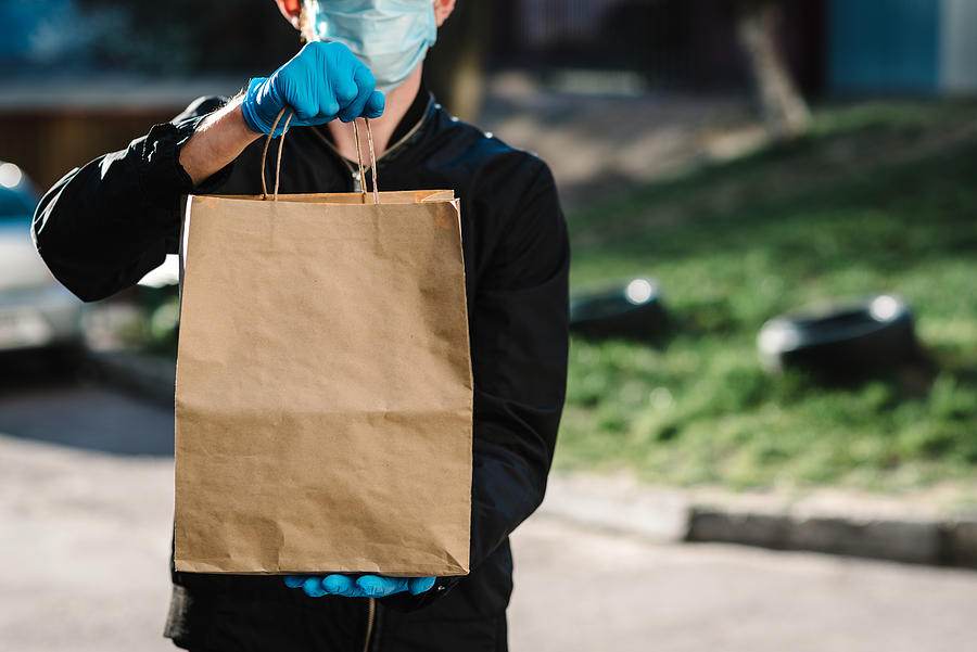 Courier in protective mask, medical gloves delivers takeaway food.  Employee hold cardboard package. Place for text. Delivery service under quarantine, 2019-ncov, pandemic coronavirus, covid-19. Photograph by Serhii Sobolevskyi