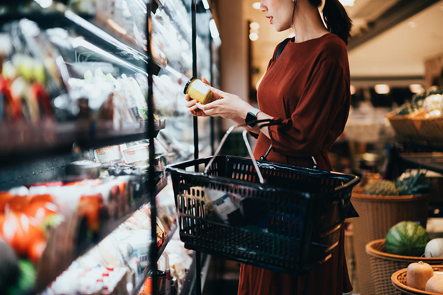 Cropped shot of young Asian woman carrying a shopping basket, standing along the dairy aisle, reading the nutrition label on the bottle of a fresh organic healthy yoghurt. Making healthier food choices Photograph by D3sign