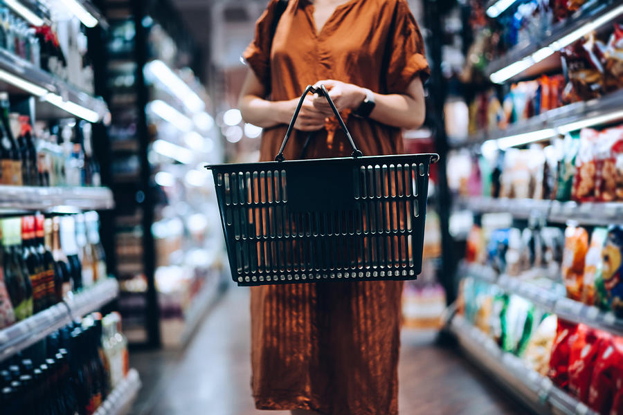 Cropped shot of young woman carrying a shopping basket, standing along the product aisle, grocery shopping for daily necessities in supermarket Photograph by D3sign