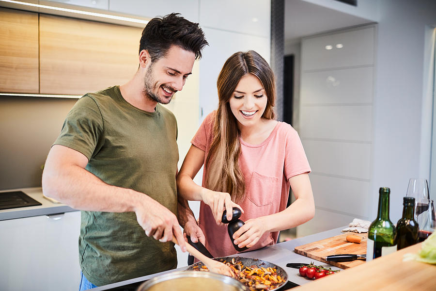 Cute joyful couple cooking together and adding spice to meal, laughing and spending time together in the kitchen Photograph by BartekSzewczyk