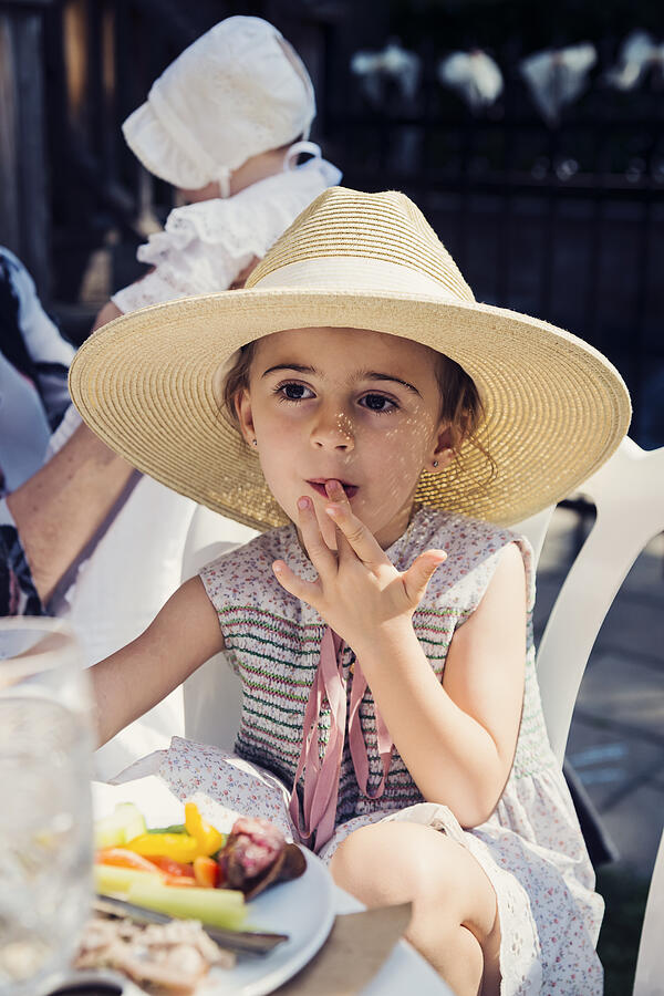 Cute little girl with too big hat eating outdoors at family reunion. Photograph by Martinedoucet