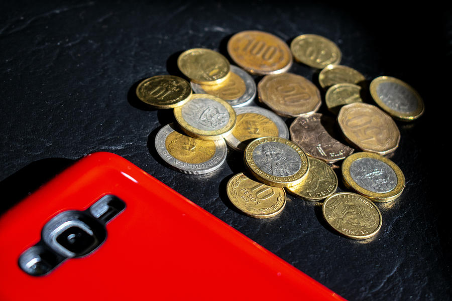 Dinero en efectivo: pesos chilenos en monedas y teléfono celular/móvil con protector rojo Photograph by Javier Ghersi