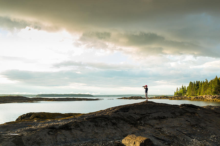 Distant view of senior man looking through binoculars at coast of Maine, USA Photograph by Heshphoto