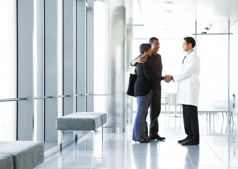 Doctor shaking hands with couple in lobby Photograph by Thomas Northcut