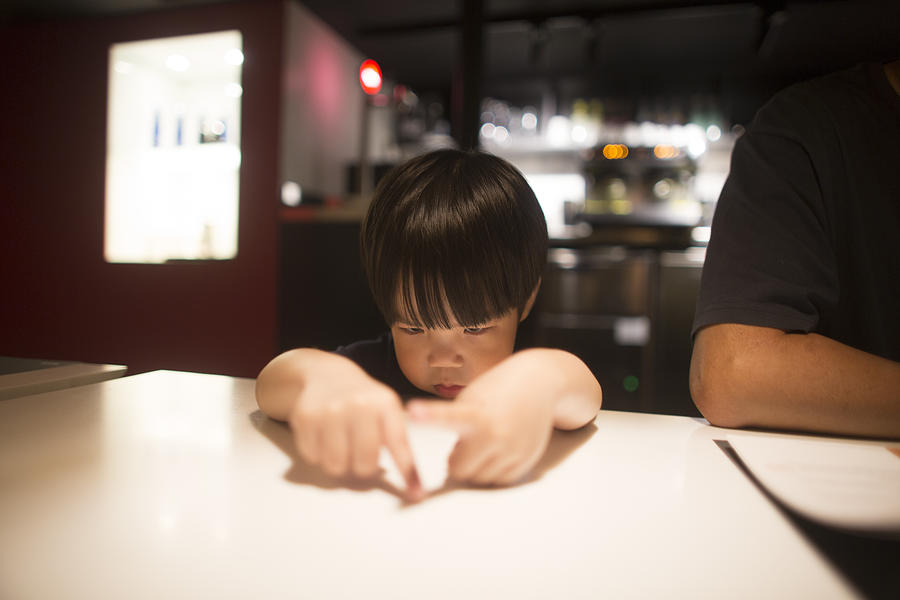 East asian young boy looking bored by waiting time in restaurant. Photograph by Twomeows