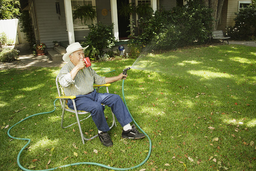 Elderly man watering lawn while sitting in chair drinking coffee Photograph by Hill Street Studios