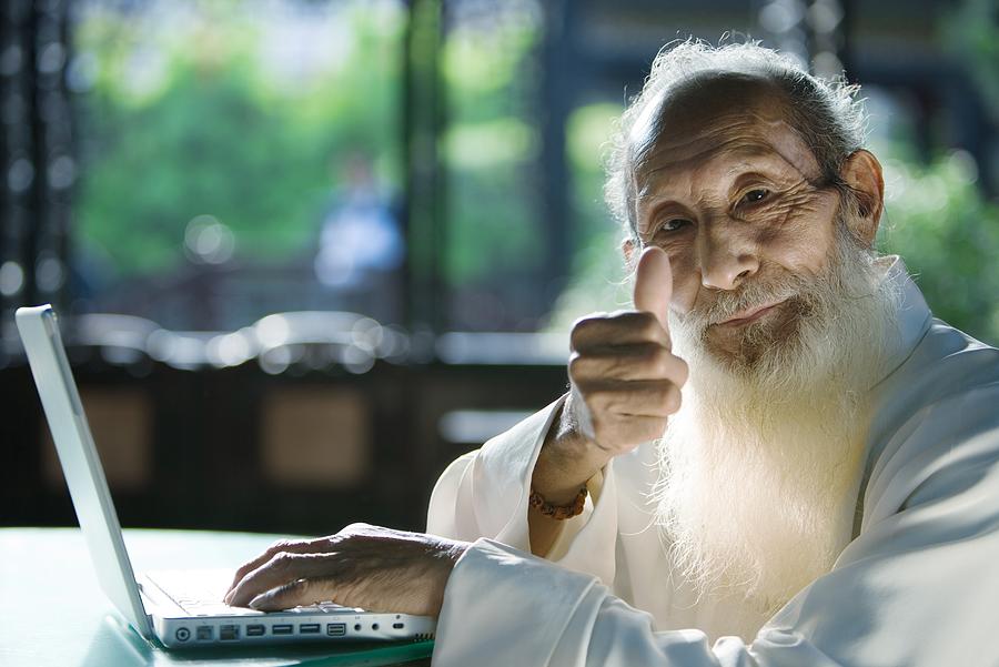 Elderly man wearing traditional Chinese clothing, using laptop, giving thumbs up to camera Photograph by ZenShui/James Hardy