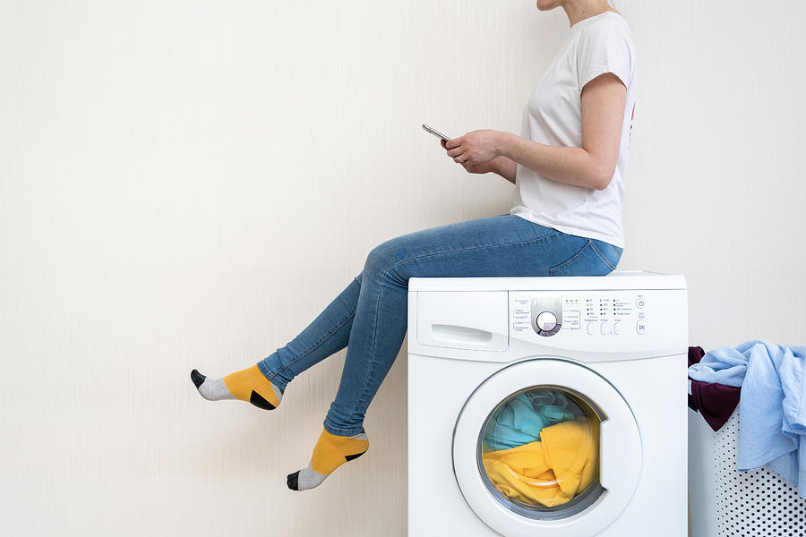 Enjoying of easy laundry process. Profile side view of lady sitting on top of white washing machine and looking at smartphone app inside bright light flat interior Photograph by Brizmaker